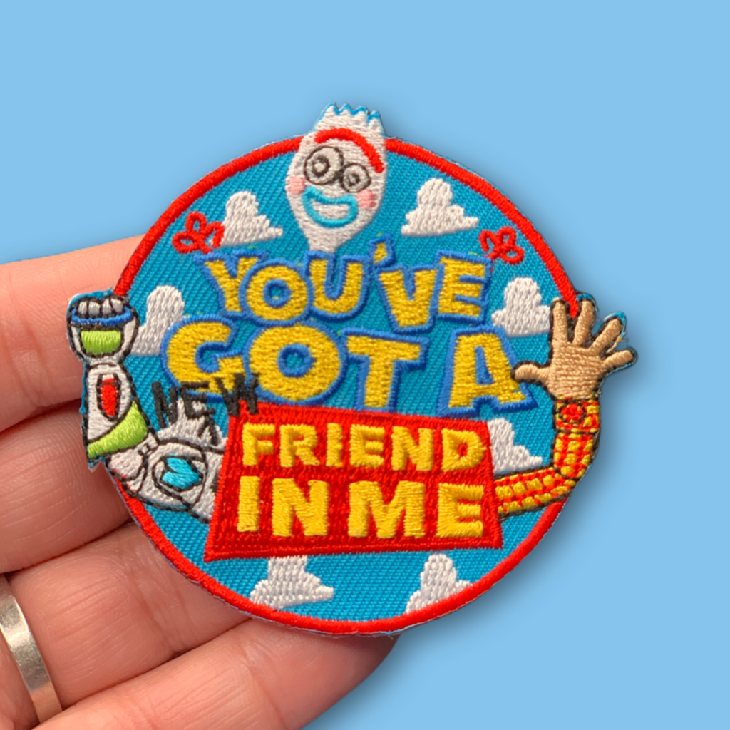 “Embroidered circular patch held in hand featuring a smiling cartoon utensil face above bold lettering, with playful illustrated arms, clouds, and bright blue, red, yellow, and white stitching.”