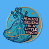 “Embroidered circular patch featuring a graceful figure in a flowing blue gown surrounded by small gold stars, with decorative lettering that reads ‘Always believe in a little magic,’ stitched in blue and gold tones.”