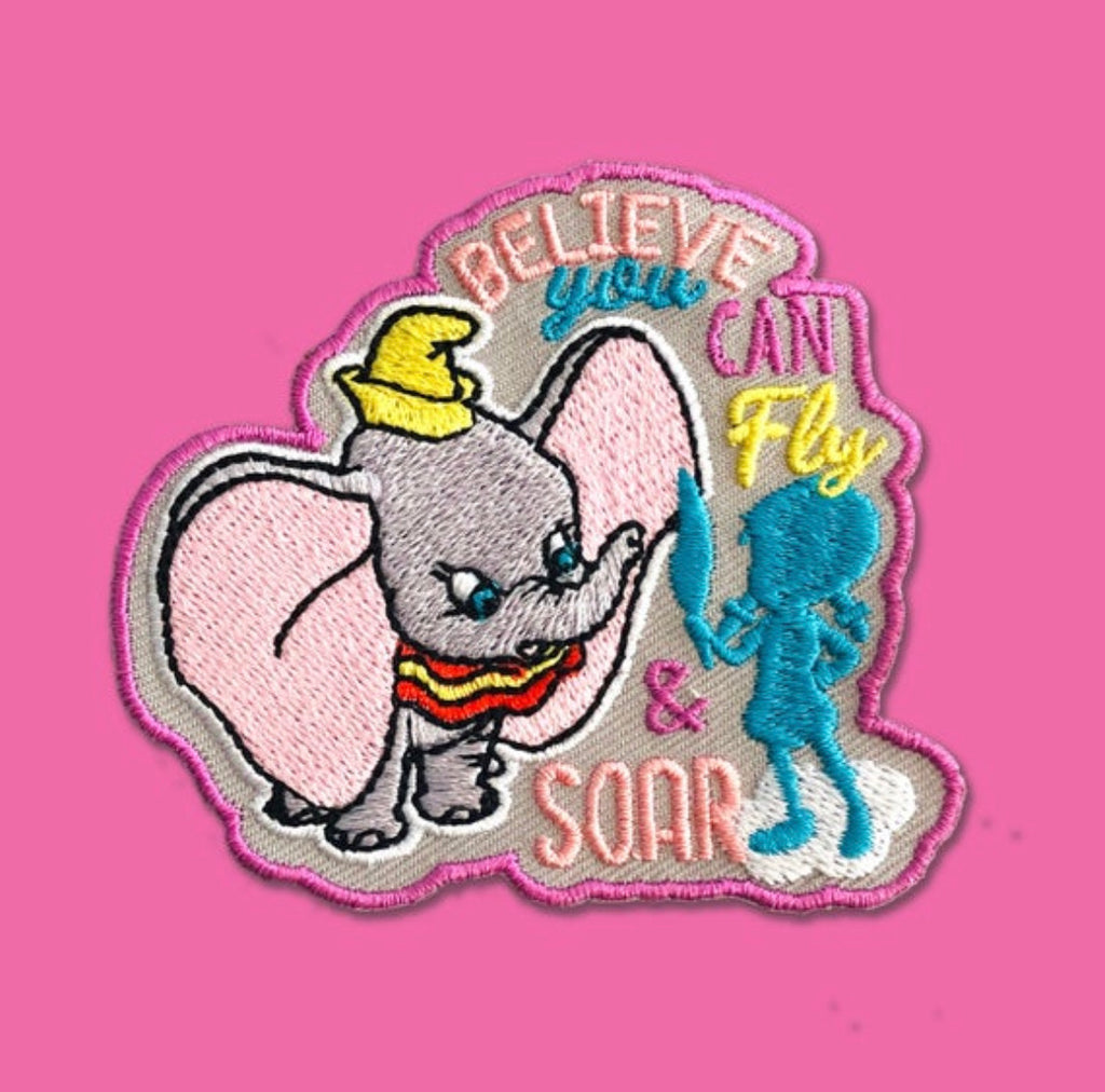 “Embroidered patch showing a smiling baby elephant wearing a small yellow hat, reaching toward a blue flying figure, with uplifting decorative lettering in pastel pink, yellow, and blue on a gray background.”