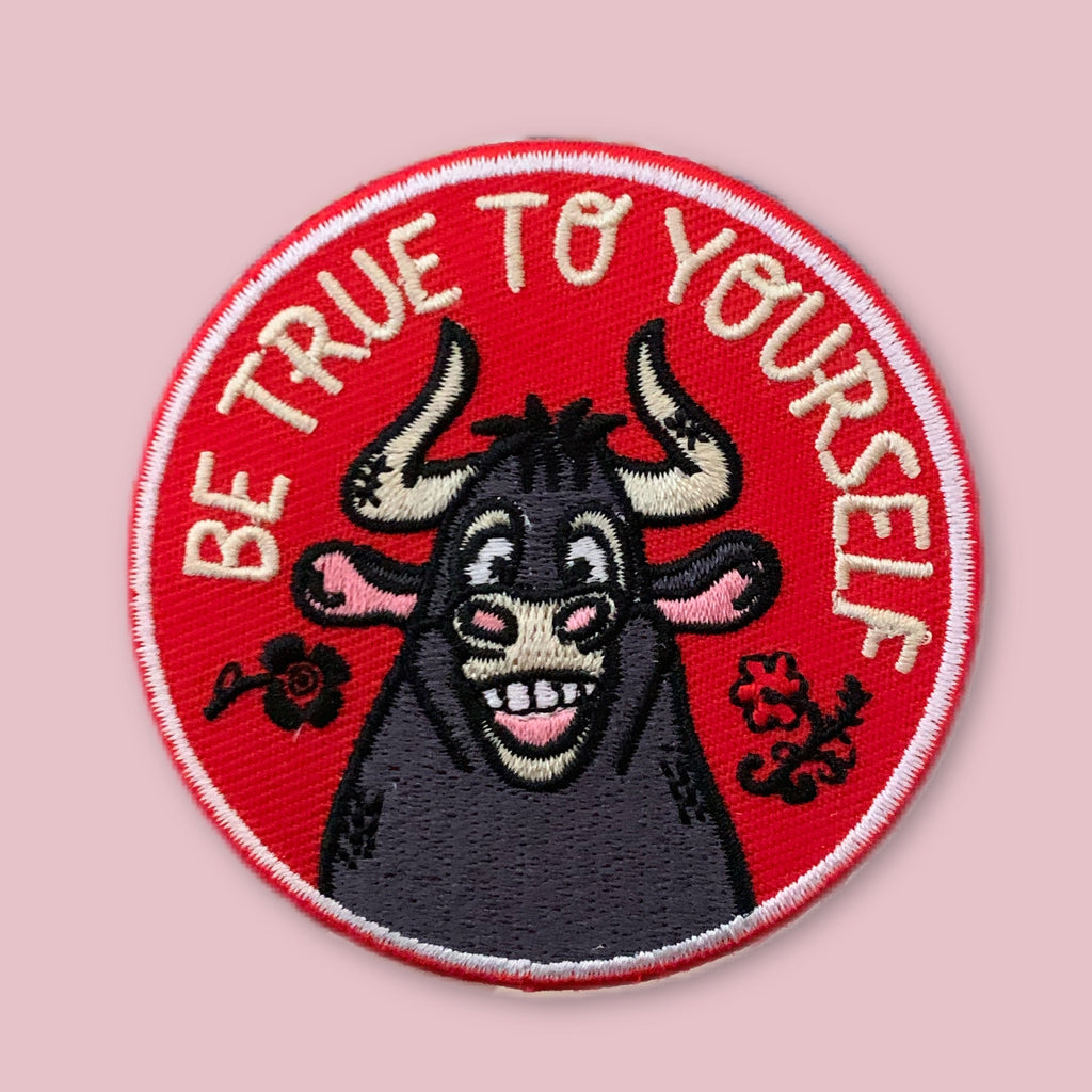 Round embroidered patch with a smiling cartoon bull on a red background, surrounded by the words “Be True to Yourself” and small floral details.