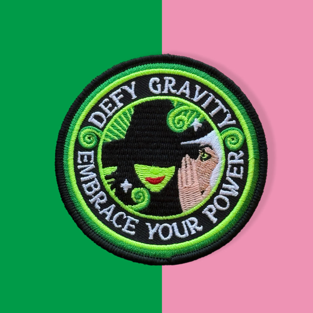 Embroidered patch featuring two stylized witch figures peeking from a green teacup with swirling steam, sparkles, a broom, and a cupcake, in bright green and pink colors on a solid background.