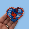 “Embroidered heart-shaped patch featuring a vintage-style figure in a blue suit and hat, split down the center, with playful stitched lettering on both sides and a scalloped red-and-white border on a blue background.”