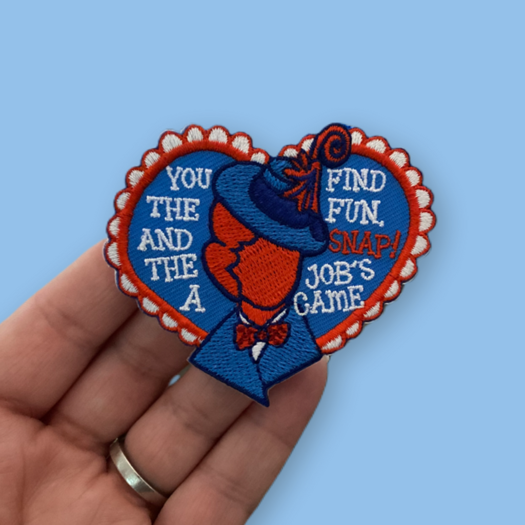 “Embroidered heart-shaped patch featuring a vintage-style figure in a blue suit and hat, split down the center, with playful stitched lettering on both sides and a scalloped red-and-white border on a blue background.”