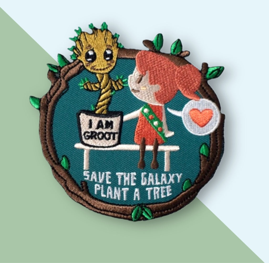 “Embroidered circular patch showing a small plant character in a flower pot sitting on a bench beside a red-haired child wearing a sash, surrounded by leafy branches, with a heart icon and stitched eco-themed message on a teal background.”