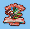 Embroidered patch showing a whimsical striped hat and colorful objects popping out of an open book, with playful shapes and bright colors on a light background.
