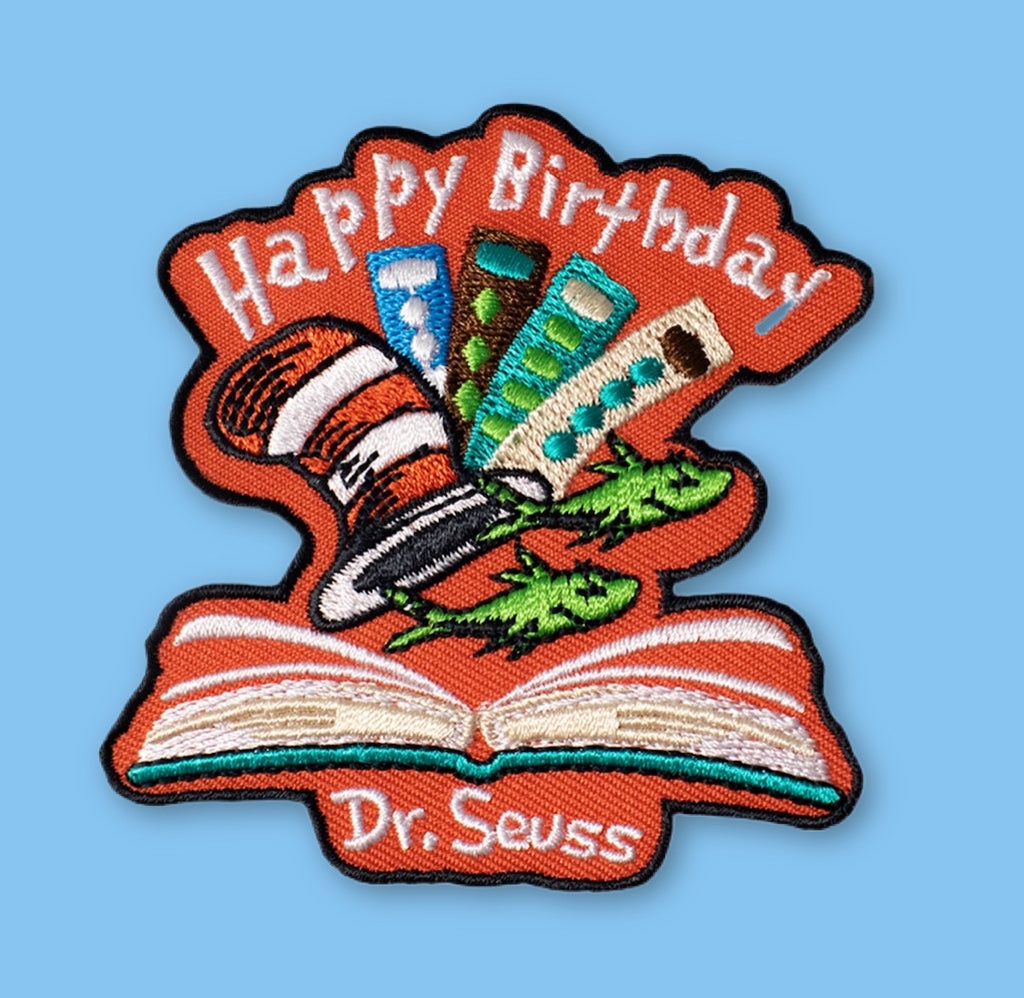 Embroidered patch showing a whimsical striped hat and colorful objects popping out of an open book, with playful shapes and bright colors on a light background.