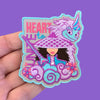 “Embroidered patch held in hand showing a stylized girl wearing a wide hat and holding a sword, surrounded by swirling clouds and flowers, with a unicorn head above, stitched in purple, teal, and pink.”