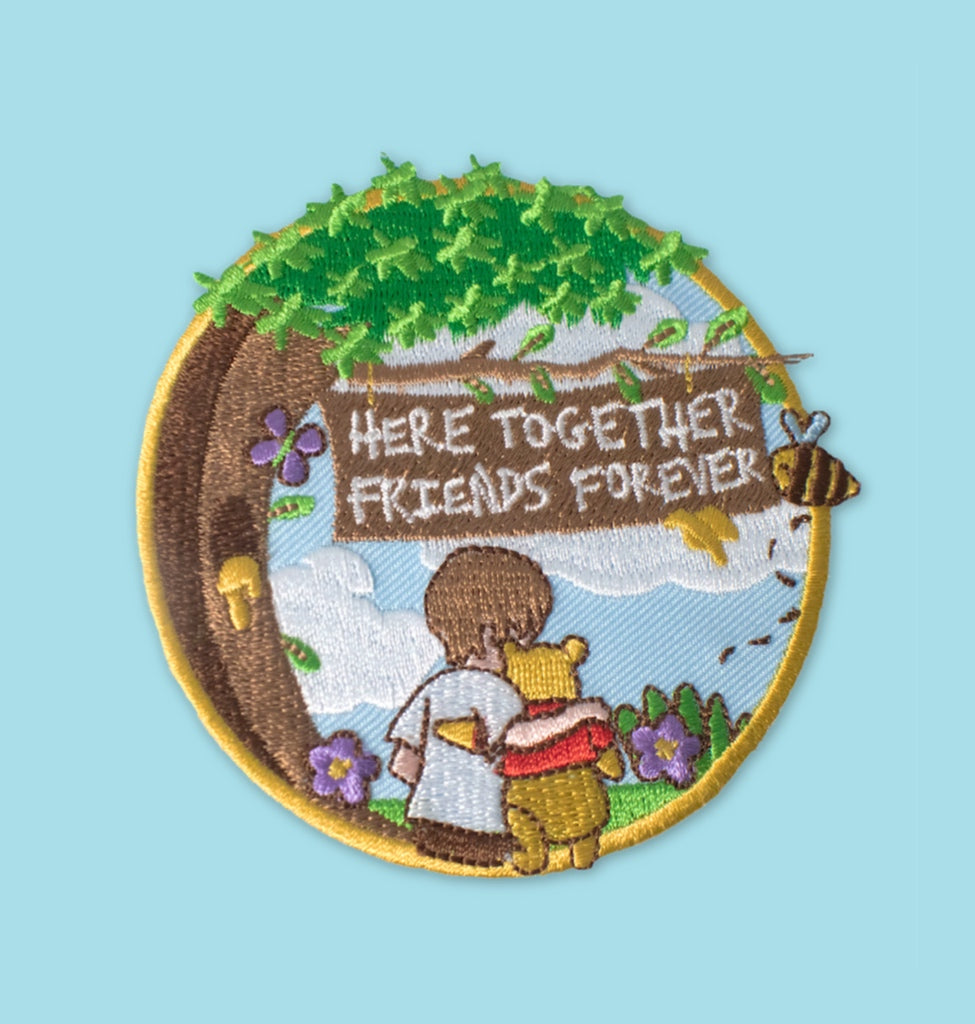 “Embroidered circular patch showing two small figures standing arm in arm beneath a leafy tree, with flowers, a bee, and a wooden sign with stitched lettering, set against a light blue sky and yellow border.”