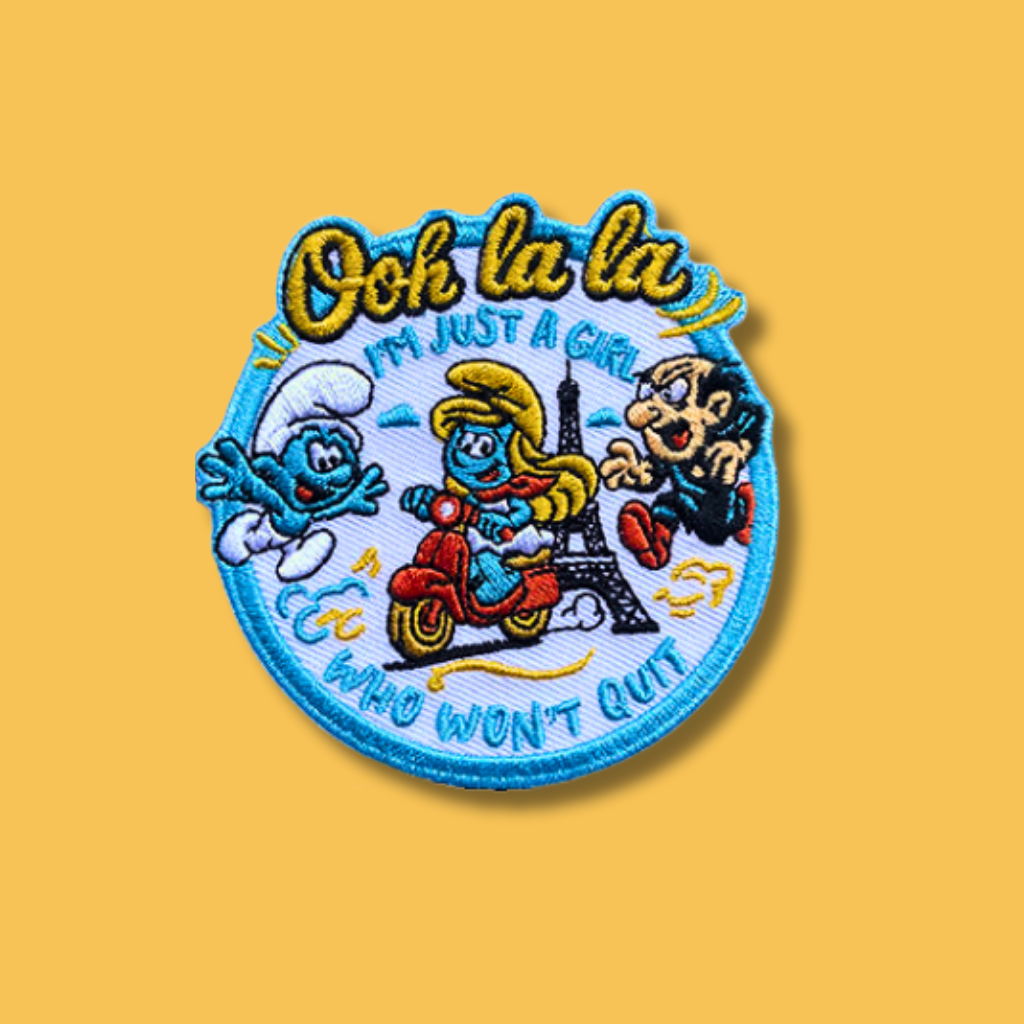 Embroidered round patch featuring blue cartoon characters riding a scooter near a city landmark with playful text.