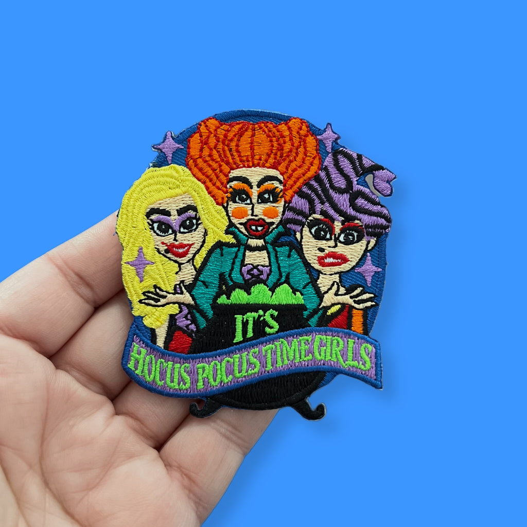 “Embroidered patch held in hand showing three stylized witch figures with colorful hair and makeup gathered around a cauldron, with decorative stars and stitched lettering.”