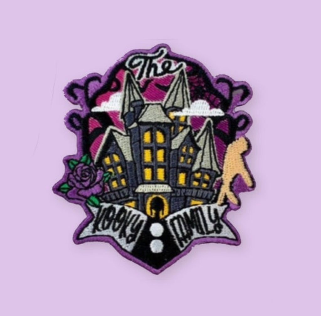 Embroidered patch featuring a dark gothic-style mansion with glowing windows, a silhouette figure nearby, decorative flowers, clouds, and a banner design in purple, black, and yellow tones.