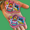 “Two embroidered rainbow cake patches displayed on a hand, each showing a stylized person holding a layered rainbow cake with colorful speech bubbles and baking details.”