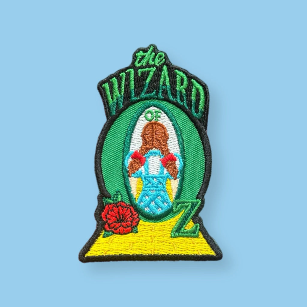 Embroidered patch featuring a stylized girl in a blue dress standing within an oval frame, with green lettering above, red flowers, and a yellow path design on a light background.