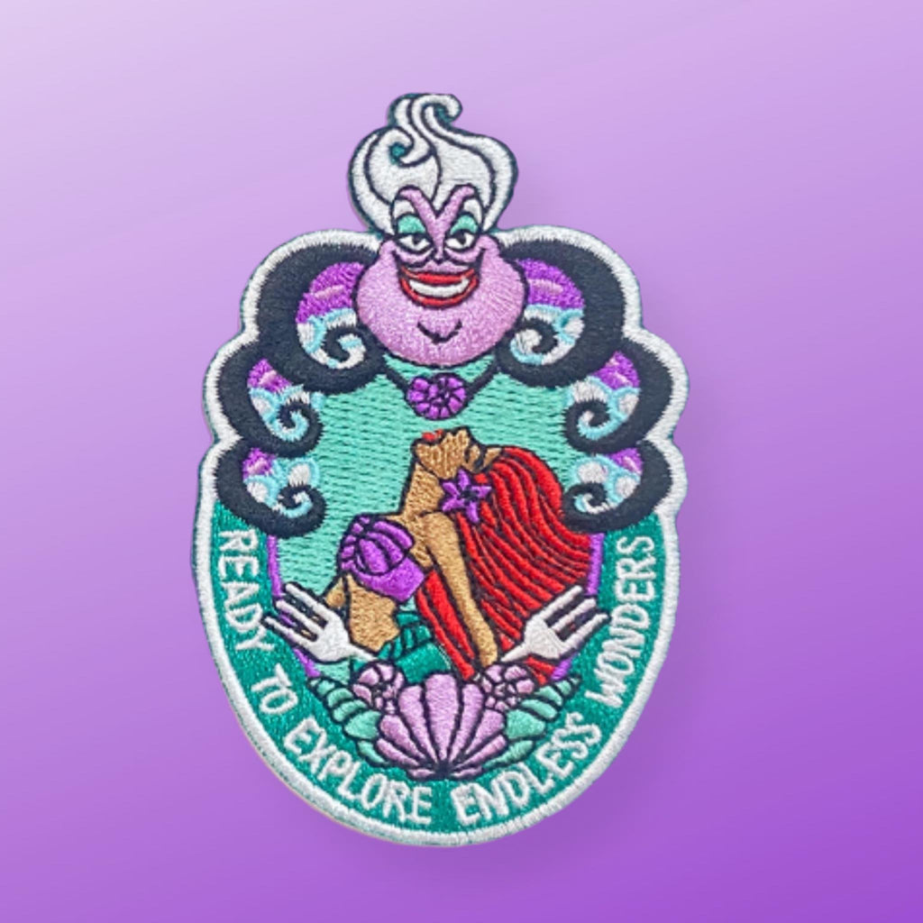 “Embroidered patch featuring a stylized sea witch figure holding a red-haired mermaid, framed by shells and flowing ocean motifs in teal, purple, and black.”