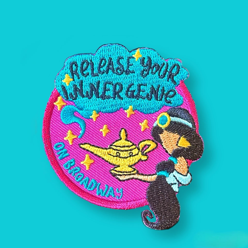 “Round embroidered patch showing a cartoon-style girl in a headband reaching toward a golden lamp, surrounded by stars and decorative stitched text in bright pink, teal, and gold.”