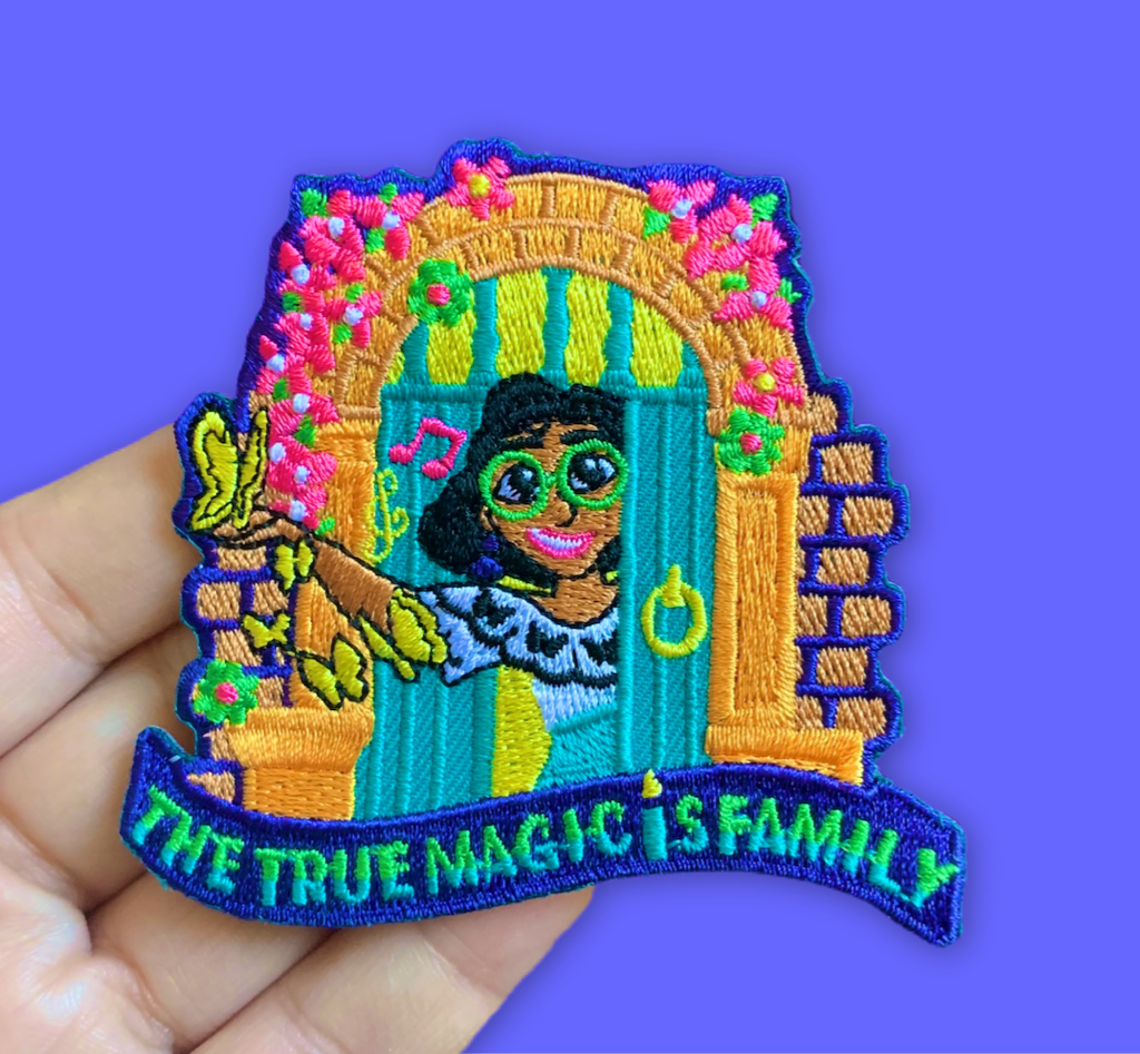 “Embroidered patch held in hand showing a smiling illustrated girl standing in a colorful doorway framed by flowers, holding butterflies, with bright teal, yellow, and pink details.”