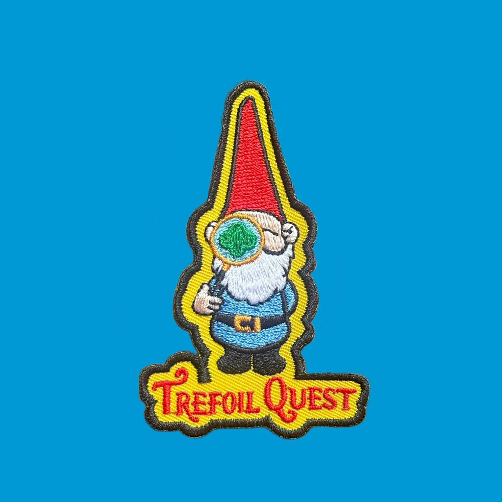 “Embroidered patch showing a small gnome with a red pointed hat and white beard holding a magnifying glass, outlined in yellow on a blue background.”