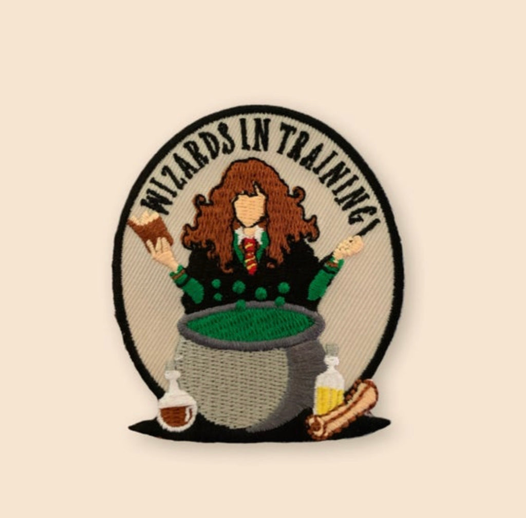 Embroidered patch showing a faceless cartoon wizard with curly hair emerging from a cauldron, holding a book and wand, with potion bottles and a wand below, and the text “Wizards in Training” on a neutral background.