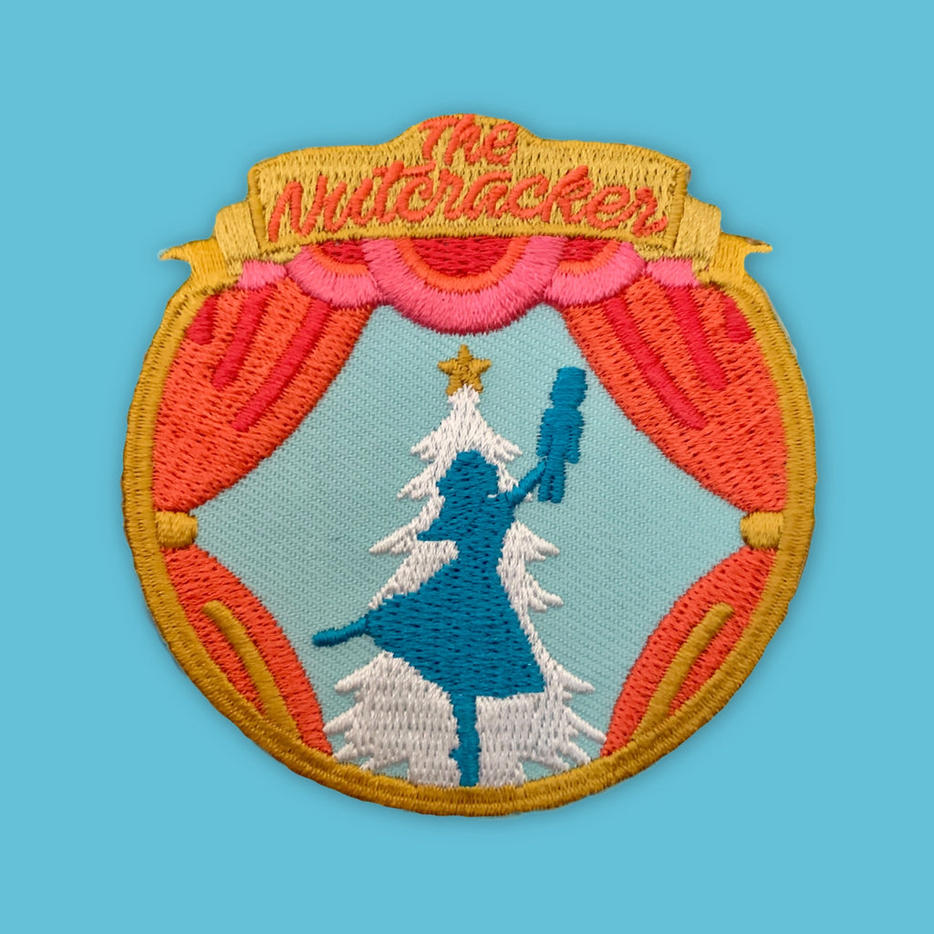 Round embroidered patch featuring a stage-like design with red curtains framing a silhouetted figure holding an object aloft, set against a wintery scene with stylized trees and a star, in warm and cool contrasting colors.