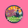 Round embroidered patch showing a joyful woman dancing in a green meadow with flowers, arms raised, set against a colorful sunrise with musical notes and decorative text elements.